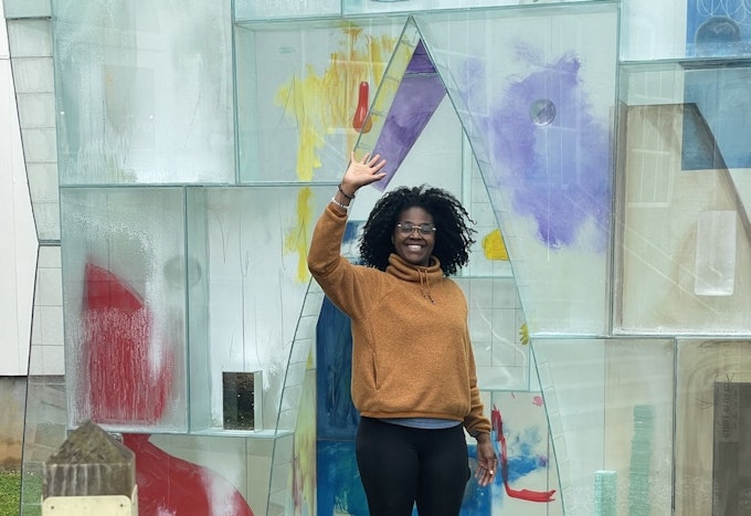 A woman in a yellow sweater smiles and waves at the camera amid a glass art sculpture outdoors.