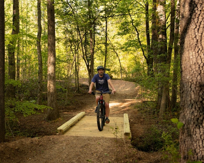 A person rides a bicycle over a wooden bridge on a forested bike path.