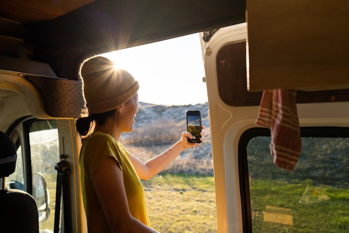 A person is standing in the doorway of a campervan holding their phone out toward the outdoors to take a photo.