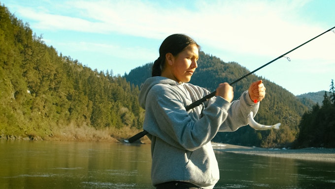 A Native American person wearing a grey hoodie is removing a fish from a fishing pole on a lake in the mountains.