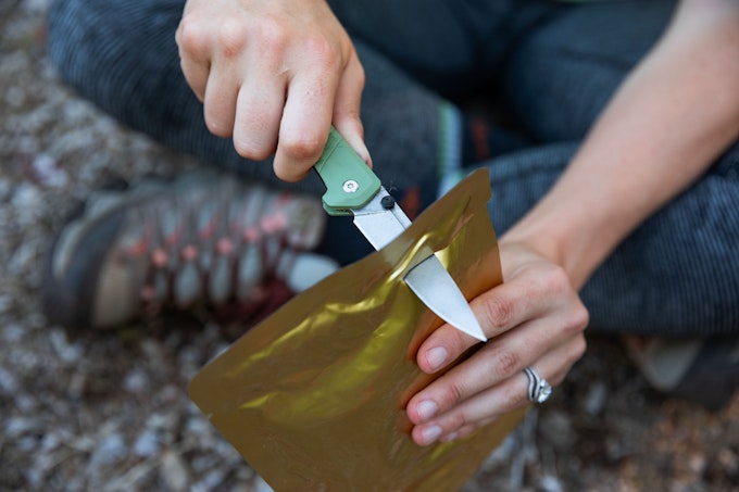 A close-up of someone using a green-handled knife to cut into a package of dehydrated backpacking food.