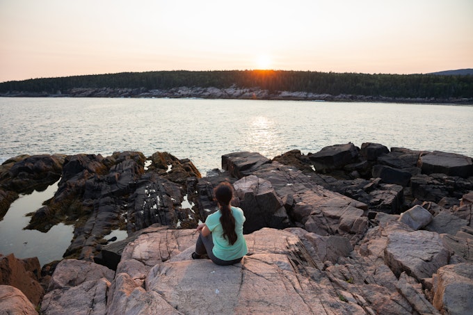 A person with a ponytail is sitting on a rocky outcropping overlooking a body of water.