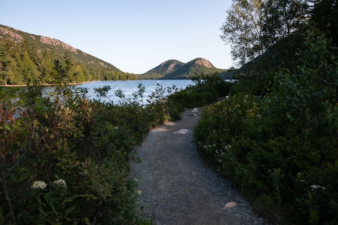 A gravel path leads away from the camera toward a lake surrounded by tree-covered mountains.