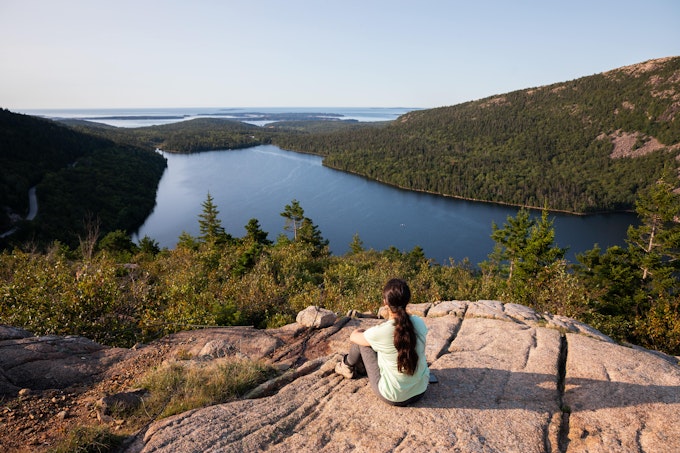 A person with a long ponytail is sitting on a rock over a lake far below. The shores are covered in pine trees and there is more water in the distance.