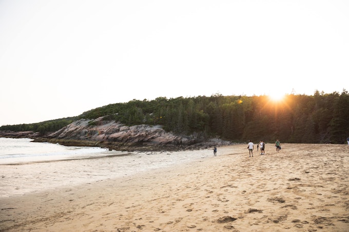 Several people are walking along a sandy beach. There is a rocky outcropping covered in trees in the background.