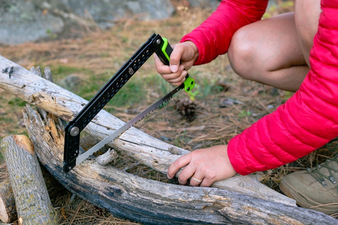 A person in shorts and a pink puffy jacket is bending down using a pack saw to saw into firewood.