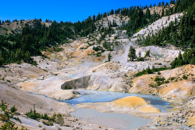 A volcanic pool is at the base of a sandy, tree-scattered hill.