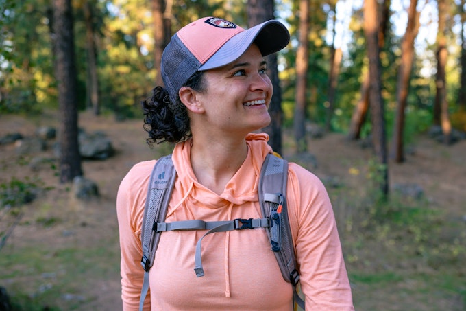 A person in a pink shirt, backpack, and pink hat is smiling off to the side. There is a multitool clipped to their backpack strap.