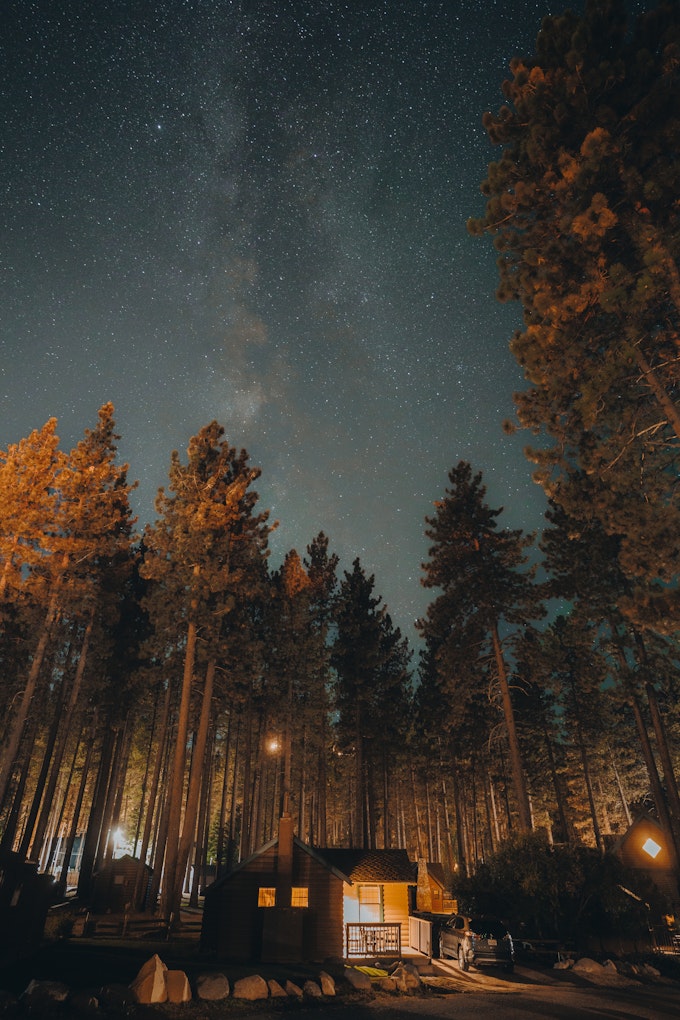 A cabin is lit up among tall pine trees and the milky way shines in the navy night sky.