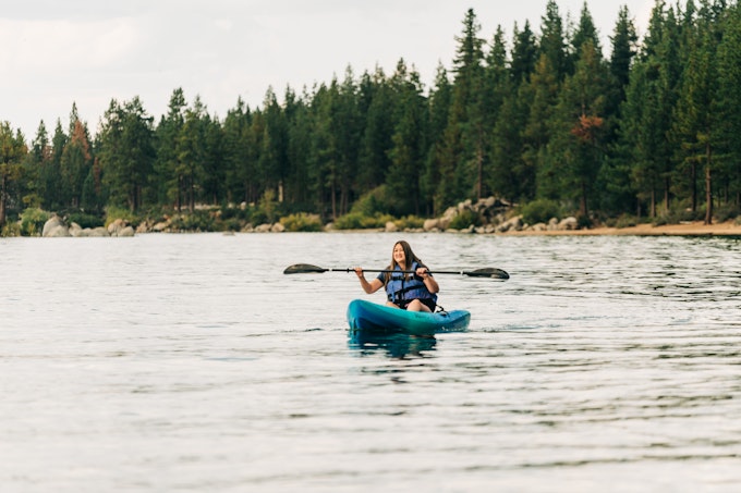 A person wearing a life jacket paddles a kayak on a body of water surrounded by trees.