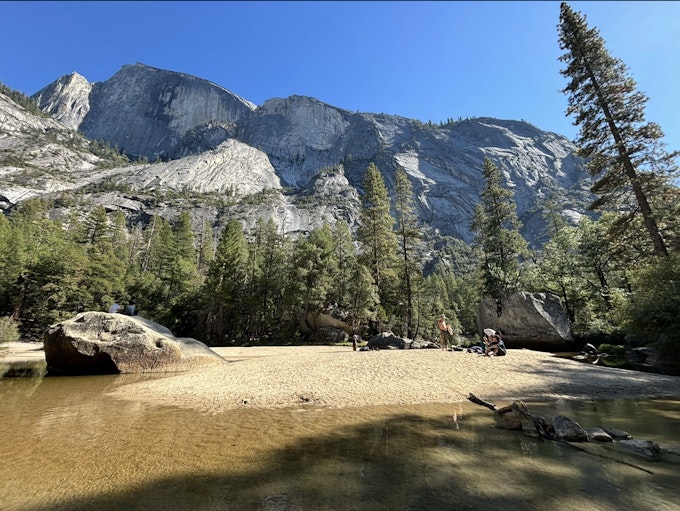 People rest on a beach under a forest and a tall, rock mountain.