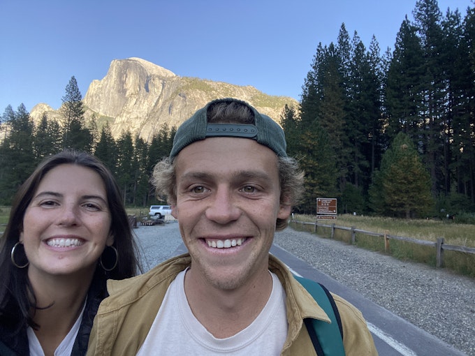 Two people are standing on a road smiling at the camera with a tall rock mountain behind them in the distance.