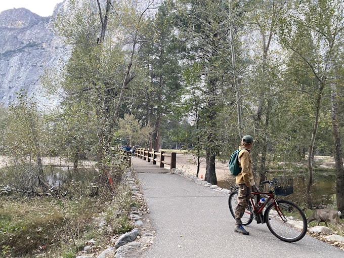 A person in a yellow jacket is stopped on their bike on a paved path near a tall, rock mountain.
