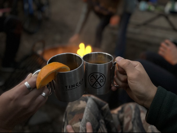 Two hands hold metal mugs to cheers over a campfire. There is an orange slice on one mug.
