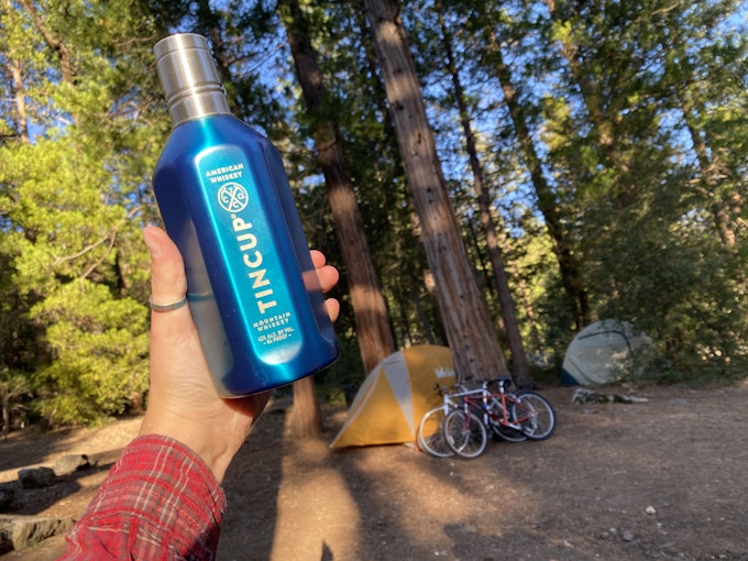 A person is holding up a metal bottle in a campground with tall trees. Bikes rest on a tree and tents are staked into the ground.