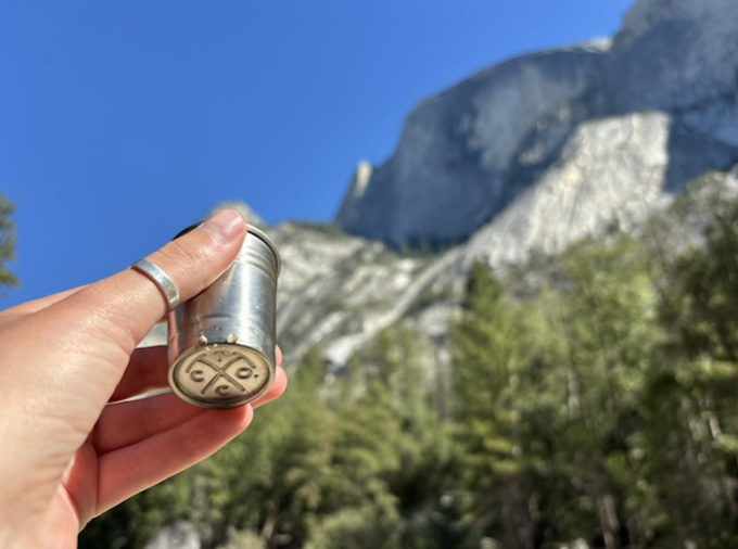 A close-up of a hand holding a small silver cup upwards toward a mountain as if cheersing it.