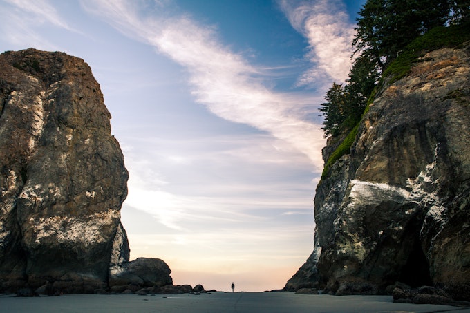 A valley of sand beach between two huge stone cliffs. A person is silhouetted far in the distance exactly in the middle of the image.