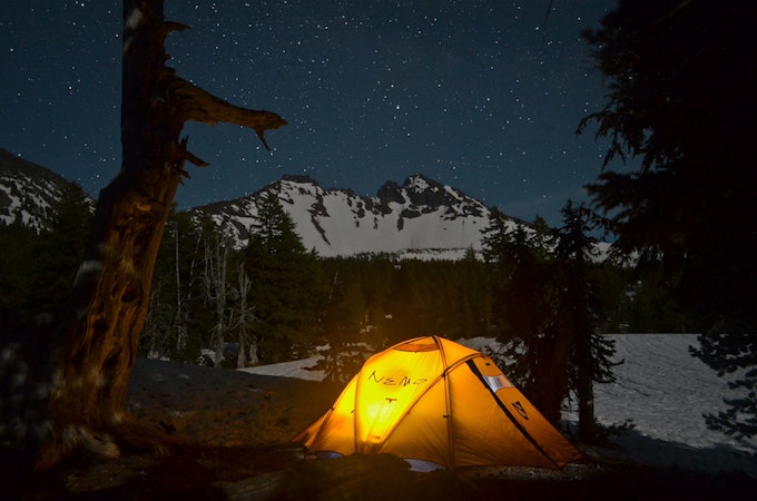 A tent glows orange in the middle of the night and is surrounded by a forest and peaks blanketed in snow