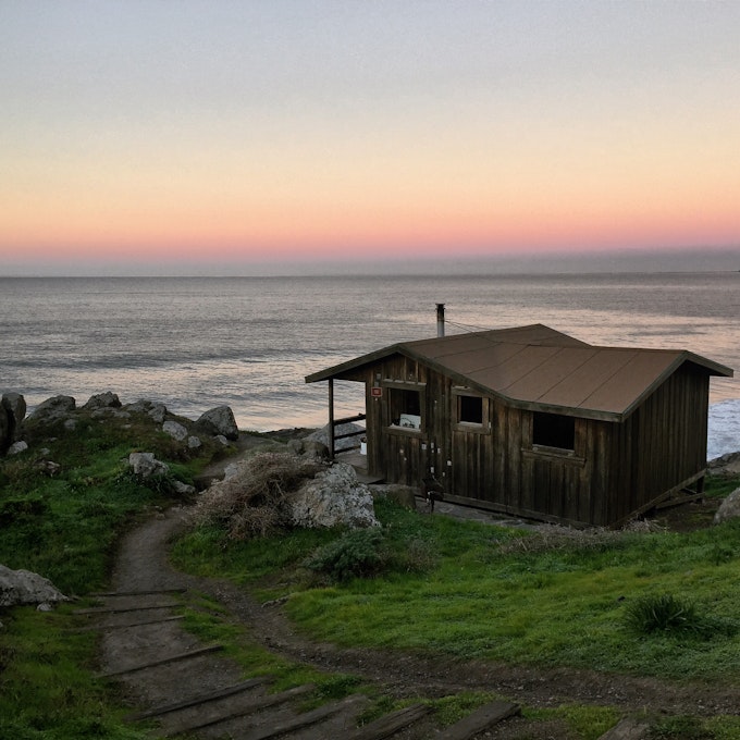 A wood building is surrounded by short grass and a large body of water is in the distance behind it.