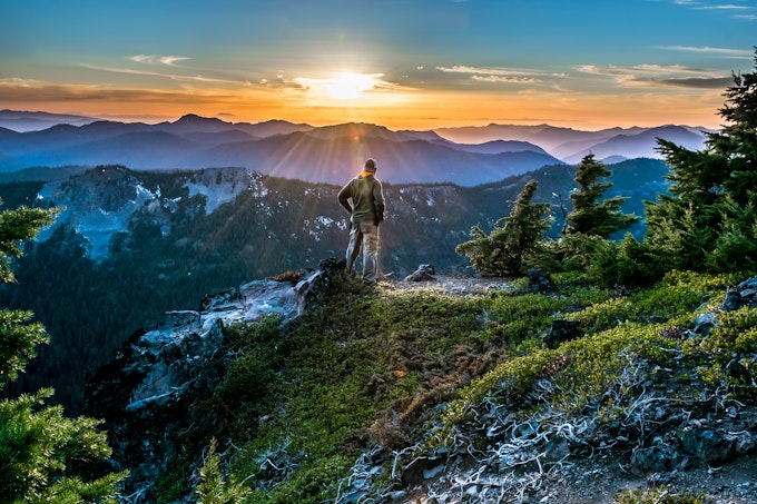 A hiker looks at the forest and mountains stretching as far as the eye can see as the sun glints over them