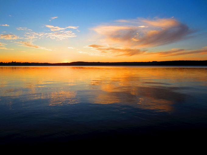 A calm lake covers the entire bottom two thirds of the image. It reflects the blue and orange sky and a few sporadic clouds.