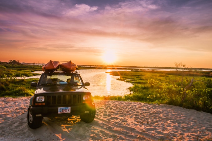 Car with two kayaks on its roof parked on the sand with a marshy background during sunset at Assateague Island.