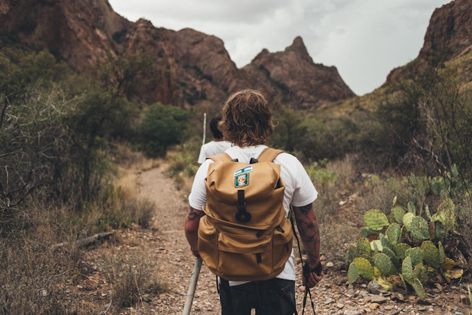 A person is wearing a yellow backpack and walking away from the camera on a Texas hike. They're holding a walking stick and there are cacti on the right side of the image. Rocky red mountains cover the background.