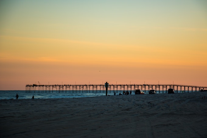 Sandy beach, ocean, and a pier in the distance during an orange sunset at Freeman Park on Carolina Beach.