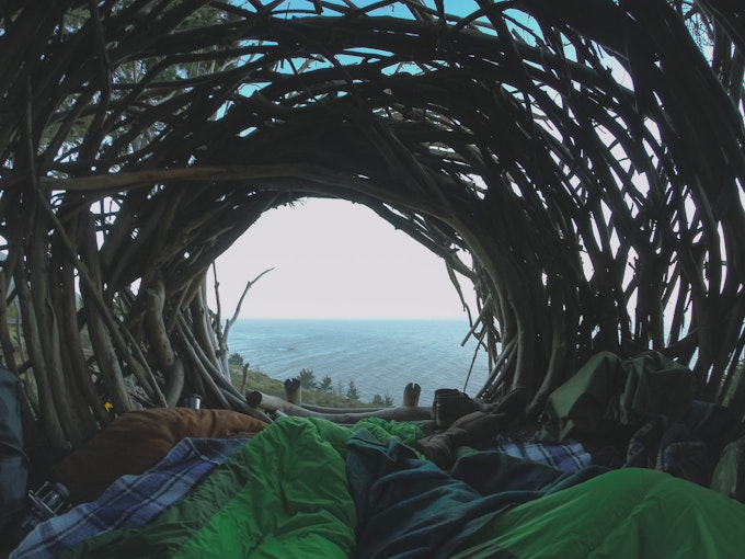 Two people are lying in sleeping bags in a massive nest-like structure looking out at a beach.