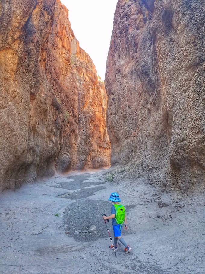 A young kid in a blue bucket hat and lime green backpack is walking on sand among tall rock walls.