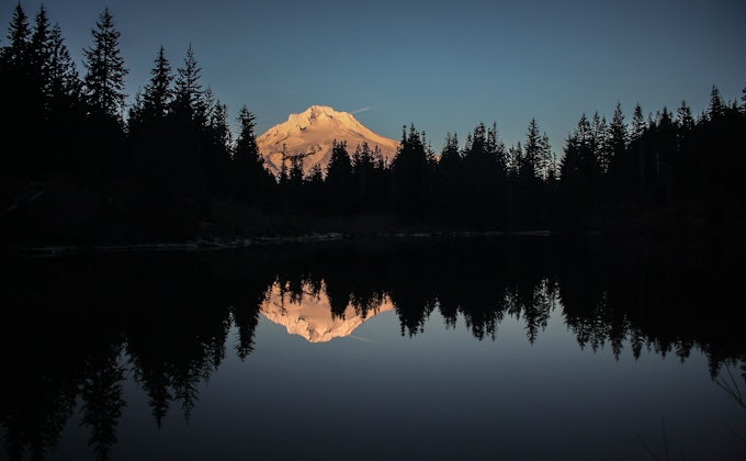 Water reflects the surrounding pine trees and snow covered mountain peak