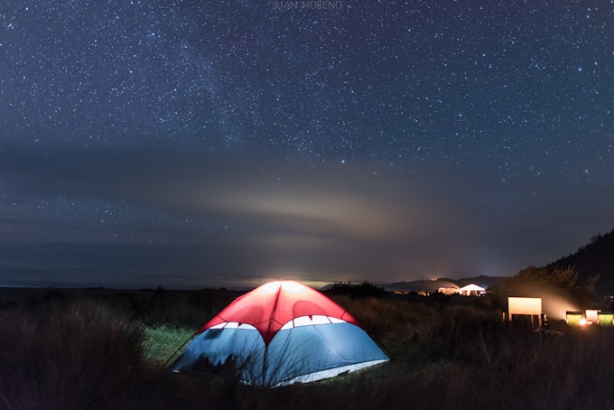 A tent is light up from inside. There is a barely visible beach in the distance and stars in the night sky.