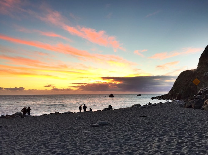 A beach at dawn or dusk with four people standing off near the water. The sky is turning orange, yellow, and pink.