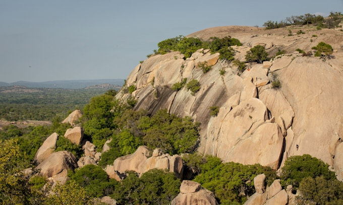 A tall, tan rock face is dotted with scrubby greenery. There's a valley in the background to the left of the rocks.