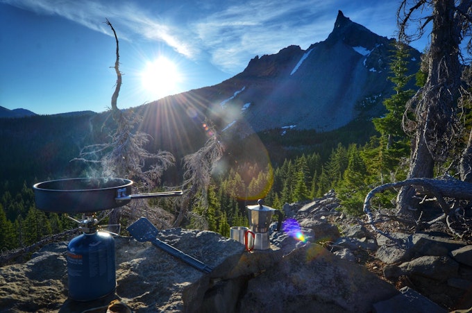 A backcountry stove sits on flat rocks with a tall, jagged peak rising in the background