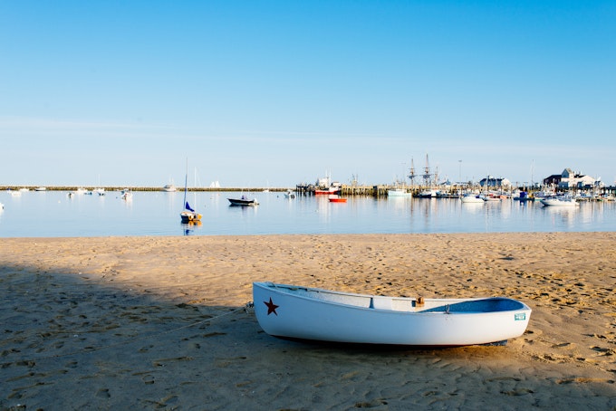 White row boat on the sand with boats floating on calm, glassy blue water in the background of Provincetown, Massachusetts.