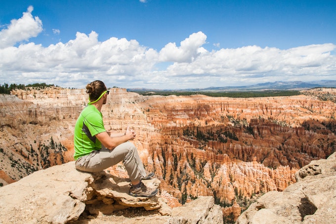 A person in khaki pants and a lime green tee is sitting on a rock overlooking red rocks. The sky is blue and puffy clouds leave shadows on the rocky cliffs.
