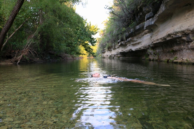 A person is soaking on a pool on a Texas hike. The water is reflecting the green plants around the edge and a rock formation with horizontal striations covers the right shoreline.