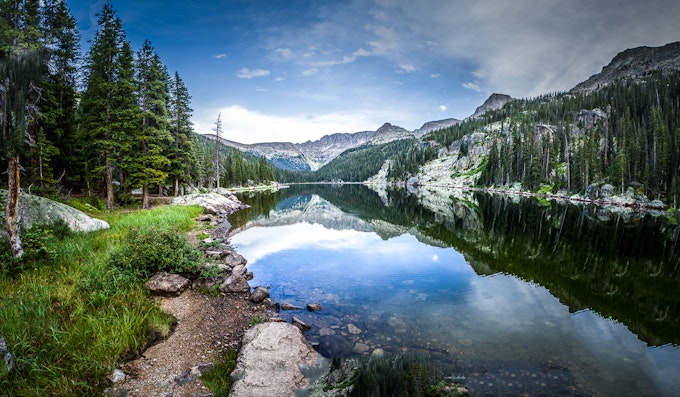A calm body of water reflect blue sky and white clouds. It is surrounded by evergreen trees and white mountain peaks.