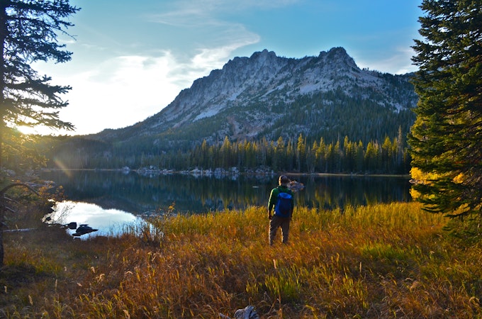 A lone hiker stands in the tan grass looking out at the lake and peak beyond
