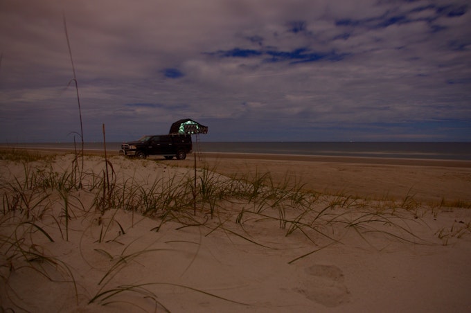 Sandy beach with some sea grass and a car with a tent setup on its roof at night. Ocean and cloudy dark sky in the background on Amelia Island.