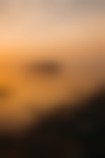 A round island rises from a large body of water. The shoreline is dotted with rocks and the water and sky has the orange tint of a sunset or sunrise.